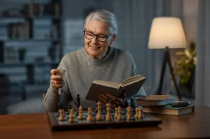 Immagine fotorealistica di una donna anziana sorridente e attiva (sui 70 anni) che gioca a scacchi o legge un libro, simboleggiando la salute cognitiva in età avanzata. Obiettivo prime 50mm, luce calda e naturale, profondità di campo per mettere a fuoco lei e sfocare leggermente lo sfondo.