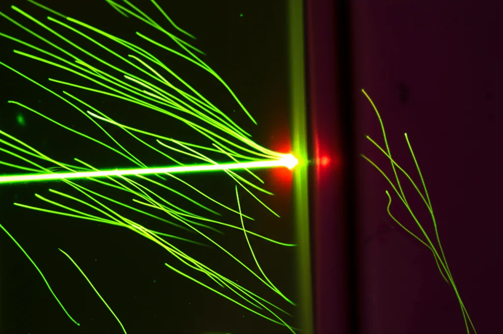 Fotografia macro di microparticelle sospese in un liquido trasparente all'interno di un dispositivo microfluidico, illuminate da un raggio laser focalizzato che crea un punto caldo visibile, con scie di flusso stilizzate che indicano il movimento. Obiettivo macro 100mm, alta definizione, illuminazione controllata, profondità di campo ridotta.