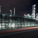 Immagine grandangolare di una moderna raffineria di petrolio di notte, obiettivo 18mm, lunga esposizione per scie luminose dalle luci e cieli lisci e scuri, focus nitido sull'intera struttura industriale illuminata con tubazioni complesse e torri, che simboleggia la complessità e l'ottimizzazione tecnologica avanzata.