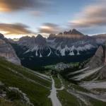 Fotografia paesaggistica grandangolare delle Dolomiti Italiane, mostrando il canalone di Cancia che scende dal Monte Antelao verso il fondovalle dove si intravedono i bacini di contenimento e il piccolo villaggio. Obiettivo grandangolare 12mm, lunga esposizione per nuvole mosse, messa a fuoco nitida su tutto il paesaggio, luce dorata del tardo pomeriggio che accentua la pendenza.