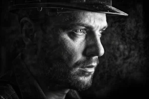 Primo piano drammatico del volto di un lavoratore industriale, illuminato lateralmente da una luce intensa che rivela particelle di polvere sospese nell'aria circostante, fotografia in bianco e nero, obiettivo 50mm, stile film noir, forte contrasto tra luci e ombre, espressione concentrata e leggermente affaticata.