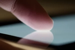 Immagine fotorealistica, obiettivo macro 100mm, che cattura un dito indice mentre interagisce con la superficie texturizzata di un moderno dispositivo touch screen (come un tablet o smartphone). Messa a fuoco nitida sulla punta del dito e sulla sottile texture del vetro. Sfondo leggermente sfocato. Alto dettaglio, illuminazione controllata che enfatizza il punto di interazione.