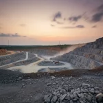 Ampia veduta paesaggistica di una cava di granito attiva in Nigeria al tramonto, macchinari pesanti visibili in lontananza, obiettivo grandangolare 18mm, esposizione lunga per catturare il movimento residuo della polvere e le nuvole soffuse, colori caldi del tramonto contrastanti con la roccia grigia e le aree scavate, messa a fuoco nitida sull'intera scena per un effetto drammatico.