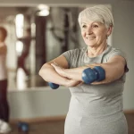 Fotografia ritratto, una donna anziana (circa 70 anni) sorridente e dall'aspetto sano, in abbigliamento sportivo, mentre si riposa un attimo durante un allenamento in una palestra per sole donne. Luce calda e motivante. Obiettivo 50mm, profondità di campo ridotta per sfocare leggermente lo sfondo della palestra. Tonalità duotone calde, tipo seppia e crema.