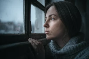 Una donna, ritratto 35mm, toni bicromatici blu e grigio, profondità di campo, che guarda fuori da una finestra in una giornata nuvolosa, trasmettendo un senso di introspezione e vulnerabilità emotiva legata all'esperienza del lutto perinatale.