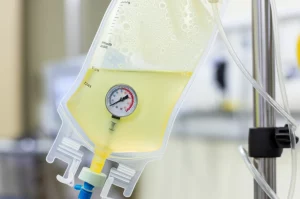 Primo piano di una sacca a pressione medica (tipo INFU-SURG) gonfia attorno a una sacca di liquido per irrigazione da 1 litro, appesa a un'asta porta flebo in un ambiente clinico luminoso. L'indicatore di pressione sulla sacca è chiaramente visibile. Obiettivo macro 90mm, alta definizione, messa a fuoco precisa sull'indicatore e sulla texture della sacca, illuminazione controllata laterale per creare profondità.