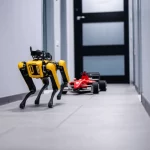 Fotografia realistica di diversi tipi di robot (un quadrupede giallo simile a Spot, uno su ruote nero come Jackal, una piccola auto da corsa F1/10 rossa) che navigano fluidamente attraverso uno stretto passaggio di porta in un ambiente interno moderno e luminoso. Obiettivo prime 35mm, profondità di campo ridotta per focalizzare l'attenzione sui robot in primo piano, illuminazione controllata e brillante, leggero effetto motion blur sulle ruote/zampe per indicare il movimento coordinato.