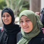 Fotografia ritratto di gruppo di diverse donne giordane di età riproduttiva, sorridenti ma con un velo di preoccupazione, ambientazione esterna luminosa, obiettivo da 50mm, profondità di campo media, colori naturali.