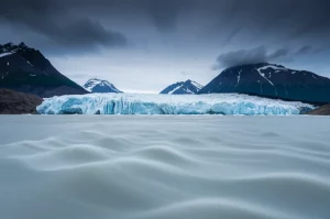 Imponente fronte di un ghiacciaio dell'Alaska che si getta in un fiume lattiginoso sottostante, montagne aspre sullo sfondo, cielo drammatico. Obiettivo grandangolare 14mm, messa a fuoco nitida su tutto il paesaggio, lunga esposizione per enfatizzare il movimento dell'acqua e delle nuvole, luce fredda del mattino.