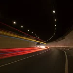 Vista grandangolare di un tunnel stradale moderno e illuminato, traffico fluido con scie luminose rosse e bianche delle auto, esposizione lunga, obiettivo 15mm, messa a fuoco nitida sull'architettura curva del tunnel, atmosfera urbana dinamica al crepuscolo.