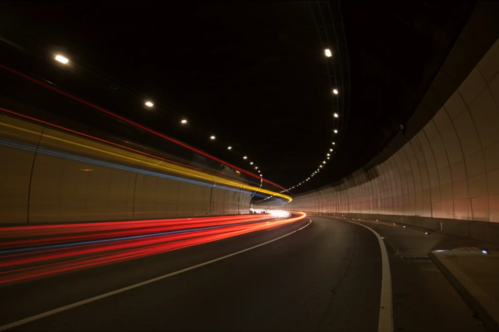 Vista grandangolare di un tunnel stradale moderno e illuminato, traffico fluido con scie luminose rosse e bianche delle auto, esposizione lunga, obiettivo 15mm, messa a fuoco nitida sull'architettura curva del tunnel, atmosfera urbana dinamica al crepuscolo.