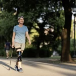 Paziente con disabilità neurologica che cammina con sicurezza e un sorriso leggero in un parco cittadino, utilizzando un discreto dispositivo FES sulla gamba, luce naturale del tardo pomeriggio che crea lunghe ombre, obiettivo prime 35mm, profondità di campo ridotta che sfoca lo sfondo verde degli alberi, mettendo in risalto la persona e il senso di libertà ritrovata.