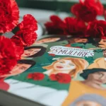 Composizione artistica fotorealistica: il libro '25 Mulheres' aperto mostra illustrazioni vivaci di donne portoghesi diverse; sullo sfondo, garofani rossi sfocati simboleggiano la Rivoluzione dei Garofani. Obiettivo 50mm, luce naturale morbida e diffusa, profondità di campo che mette a fuoco il libro ma lascia intravedere i fiori, colori saturi ma realistici.