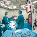 Immagine fotorealistica di una sala operatoria moderna durante un intervento di nefrectomia laparoscopica, vista grandangolare 18mm che mostra il team chirurgico composto da 3 persone concentrate sui monitor e sul paziente, luci chirurgiche brillanti ma focalizzate sull'area operatoria, sharp focus su tutta la scena.