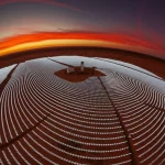 Imponente torre solare a concentrazione (CSP) circondata da eliostati scintillanti nel vasto deserto algerino, ripresa con obiettivo grandangolare 18mm al tramonto, cielo infuocato, alta definizione, messa a fuoco nitida sull'intera scena.