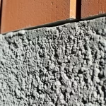 Immagine fotorealistica di un moderno edificio urbano in costruzione, con primo piano su una sezione di muro in calcestruzzo ecologico realizzato con scorie d'acciaio al posto degli inerti naturali. Dettagli macro sulla texture porosa ma compatta del calcestruzzo. Obiettivo grandangolare 24mm per catturare la scala urbana e l'innovazione materica, luce naturale.