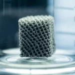 Immagine fotorealistica, scattata con un obiettivo macro da 100mm, che mostra un sofisticato elettrodo di carbonio poroso stampato in 3D, di colore grigio scuro, con una complessa struttura a reticolo. L'elettrodo è parzialmente immerso in un elettrolita trasparente all'interno di un beaker di vetro, con sottili riflessi di luce sulla superficie. Alto dettaglio, illuminazione controllata da laboratorio.