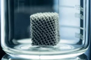 Immagine fotorealistica, scattata con un obiettivo macro da 100mm, che mostra un sofisticato elettrodo di carbonio poroso stampato in 3D, di colore grigio scuro, con una complessa struttura a reticolo. L'elettrodo è parzialmente immerso in un elettrolita trasparente all'interno di un beaker di vetro, con sottili riflessi di luce sulla superficie. Alto dettaglio, illuminazione controllata da laboratorio.