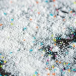 Immagine fotorealistica macro, obiettivo 105mm, alta definizione, illuminazione controllata da studio, che mostra polvere bianca fine di chitosano che sembra intrappolare o aggregare piccole particelle colorate di microplastica su uno sfondo neutro.