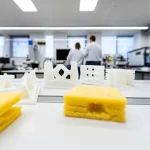 Scatto grandangolare, obiettivo 24mm, che mostra diverse strutture sandwich in schiuma di PLA stampate in 3D con diverse densità e pattern interni, disposte esteticamente. Alcune mostrano danni da impatto. Ambiente di laboratorio luminoso e pulito, che enfatizza innovazione e sostenibilità.