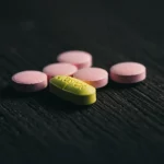 Primo piano di diverse compresse di rosuvastatina, alcune generiche rosa e una di marca (Crestor®) gialla, disposte artisticamente su una superficie di legno scuro, obiettivo macro 90mm, profondità di campo ridotta, illuminazione da studio laterale che crea ombre morbide.