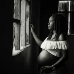 Fotografia di ritratto toccante di una donna Ghanese incinta che guarda fuori da una finestra nella Volta Region, Ghana. Luce soffusa, obiettivo 50mm, profondità di campo che isola la figura, espressione di speranza mista a preoccupazione. Bianco e nero cinematografico, stile film noir.