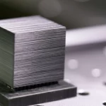Immagine fotorealistica di un campione cubico di Invar-TiCN stampato in 3D (aspetto metallico grigio scuro con texture stratificata fine) posizionato su un banco da laboratorio accanto a un diffrattometro a neutroni stilizzato, obiettivo prime 35mm, profondità di campo ridotta, illuminazione da laboratorio controllata e focalizzata sul campione.