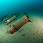 Immagine subacquea drammatica e fotorealistica di diverse munizioni della Seconda Guerra Mondiale sparse sul fondale marino tedesco, alcune parzialmente sepolte nella sabbia, luce filtrante dall'alto, obiettivo grandangolare 20mm, messa a fuoco nitida sui dettagli corrosi.