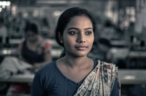 Ritratto intenso di una giovane donna bengalese, operaia tessile, sguardo pensieroso ma resiliente, ambientazione sfocata di una fabbrica tessile. Fotografia stile ritratto, obiettivo 35mm, toni seppia e blu duotone, profondità di campo.