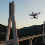 Drone in volo stazionario ispeziona da vicino la struttura complessa di un pilone di ponte in costruzione, in un ambiente montuoso al tramonto. Obiettivo zoom 35mm, profondità di campo per mettere a fuoco sia il drone che i dettagli del ponte, luce calda del tramonto.
