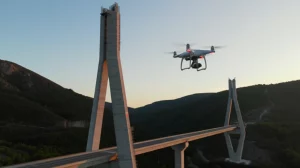 Drone in volo stazionario ispeziona da vicino la struttura complessa di un pilone di ponte in costruzione, in un ambiente montuoso al tramonto. Obiettivo zoom 35mm, profondità di campo per mettere a fuoco sia il drone che i dettagli del ponte, luce calda del tramonto.