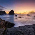 Immagine grandangolare di una costa frastagliata dell'Oregon al tramonto, obiettivo 15mm, lunga esposizione per rendere l'acqua setosa, messa a fuoco nitida sulle rocce in primo piano, cielo con colori caldi, simboleggia la bellezza minacciata dal cambiamento climatico e la necessità di azione collettiva basata sull'efficacia relazionale.