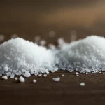 Primo piano di un mucchietto di sale bianco normale accanto a un mucchietto di sostituto del sale (leggermente più opaco e granuloso) su una superficie di legno scuro. Luce naturale laterale che crea ombre morbide, obiettivo macro 90mm, alta definizione dei cristalli, sfondo sfocato che suggerisce un ambiente di cucina salutare.