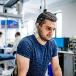 Fotografia realistica di un uomo con lesione midollare cervicale che sta in piedi con successo utilizzando la stimolazione epidurale spinale e un deambulatore per l'equilibrio in un ambiente di ricerca clinica avanzato. L'uomo mostra un'espressione di sforzo concentrato e determinazione. L'attrezzatura di stimolazione (fili sottili sulla schiena collegati a un'unità esterna) è visibile. L'illuminazione è controllata, tipica di un laboratorio, mettendo in risalto il soggetto. Obiettivo prime 50mm, profondità di campo ridotta per isolare il soggetto dallo sfondo leggermente sfocato dove si intravedono ricercatori e attrezzature. Stile fotogiornalistico scientifico.