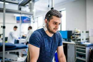 Fotografia realistica di un uomo con lesione midollare cervicale che sta in piedi con successo utilizzando la stimolazione epidurale spinale e un deambulatore per l'equilibrio in un ambiente di ricerca clinica avanzato. L'uomo mostra un'espressione di sforzo concentrato e determinazione. L'attrezzatura di stimolazione (fili sottili sulla schiena collegati a un'unità esterna) è visibile. L'illuminazione è controllata, tipica di un laboratorio, mettendo in risalto il soggetto. Obiettivo prime 50mm, profondità di campo ridotta per isolare il soggetto dallo sfondo leggermente sfocato dove si intravedono ricercatori e attrezzature. Stile fotogiornalistico scientifico.