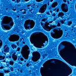 Immagine macrofotografica ad alta definizione di cellule staminali mesenchimali su uno scaffold biocompatibile tridimensionale, illuminate da una luce bluastra che simboleggia l'attivazione genica tramite mRNA per la rigenerazione ossea. Obiettivo macro 100mm, illuminazione controllata da laboratorio, messa a fuoco precisa sui dettagli cellulari e sulla microstruttura porosa dello scaffold, sfondo scuro per enfatizzare il soggetto.