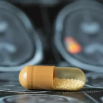 Primo piano di una capsula di meldonium su uno sfondo sfocato di una risonanza magnetica cerebrale con un glioblastoma evidenziato. Illuminazione da laboratorio controllata, lente macro 90mm, alta definizione, focus preciso sulla capsula.