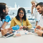 Un gruppo eterogeneo di studenti di odontoiatria collabora attivamente attorno a un tavolo in un laboratorio moderno, utilizzando modelli dentali e tablet. Un docente sorridente osserva in background. Fotografia con obiettivo prime 35mm, luce naturale morbida, profondità di campo che sfoca leggermente lo sfondo per concentrarsi sul gruppo.