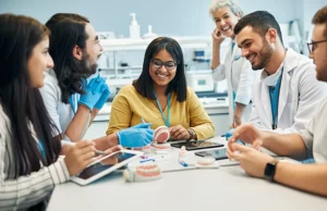 Un gruppo eterogeneo di studenti di odontoiatria collabora attivamente attorno a un tavolo in un laboratorio moderno, utilizzando modelli dentali e tablet. Un docente sorridente osserva in background. Fotografia con obiettivo prime 35mm, luce naturale morbida, profondità di campo che sfoca leggermente lo sfondo per concentrarsi sul gruppo.