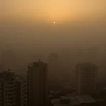 Veduta aerea di Bukan, Iran, avvolta da una densa foschia da inquinamento atmosferico al tramonto. Wide-angle lens 15mm, long exposure per sfumare le luci della città e accentuare la cappa di smog, sharp focus sugli edifici più alti che emergono dalla foschia, colori caldi ma cupi.