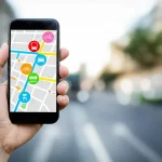 Immagine fotorealistica di un'interfaccia utente di un'app MaaS (Mobility-as-a-Service) visualizzata su uno smartphone tenuto in mano. L'app mostra una mappa cittadina stilizzata con icone che rappresentano diverse opzioni di trasporto disponibili: un autobus, un tram, una bicicletta (bike sharing) e un'auto (car sharing). Catturata con un obiettivo standard da 50mm che crea una piacevole profondità di campo sfocando leggermente lo sfondo urbano. I colori dell'interfaccia sono vivaci e moderni.