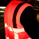 Fotografia di un ciclista di notte che indossa un gilet ad alta visibilità in tessuto misto poliestere/bambù tinto con colorante fluorescente rosso, il gilet brilla intensamente sotto i fari di un'auto. Scatto d'azione con teleobiettivo 100mm, fast shutter speed per catturare il movimento, effetto movimento leggero sullo sfondo urbano notturno, focus nitido sul tessuto brillante.