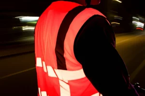 Fotografia di un ciclista di notte che indossa un gilet ad alta visibilità in tessuto misto poliestere/bambù tinto con colorante fluorescente rosso, il gilet brilla intensamente sotto i fari di un'auto. Scatto d'azione con teleobiettivo 100mm, fast shutter speed per catturare il movimento, effetto movimento leggero sullo sfondo urbano notturno, focus nitido sul tessuto brillante.
