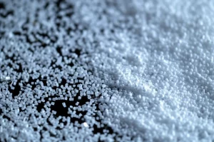 Primo piano macro di particelle di PMMA riciclato di diverse dimensioni, alcune fini altre più grandi, illuminate con luce da laboratorio controllata, messa a fuoco precisa sui dettagli delle particelle, obiettivo macro 100mm, alta definizione.