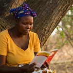 Ritratto di una donna del Botswana, seduta all'aperto sotto un albero, che legge con attenzione un opuscolo colorato 'Living positive with HIV'. Obiettivo prime 35mm, profondità di campo ridotta per sfocare lo sfondo di vegetazione locale, luce naturale calda del tardo pomeriggio, espressione concentrata ma serena e speranzosa.
