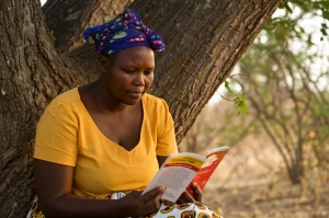 Ritratto di una donna del Botswana, seduta all'aperto sotto un albero, che legge con attenzione un opuscolo colorato 'Living positive with HIV'. Obiettivo prime 35mm, profondità di campo ridotta per sfocare lo sfondo di vegetazione locale, luce naturale calda del tardo pomeriggio, espressione concentrata ma serena e speranzosa.