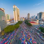 Veduta aerea dinamica del mercato di Tanah Abang oggi, che mostra il contrasto tra i moderni blocchi commerciali multipiano e il brulicare di attività nelle strade circostanti, tessuti colorati visibili sulle bancarelle esterne, luce del tardo pomeriggio che crea lunghe ombre, obiettivo grandangolare 20mm, alta definizione, tracciamento del movimento per catturare il flusso di persone e veicoli, stile fotografia urbana.