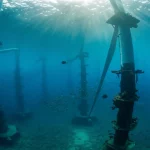 Veduta subacquea ampia di un parco eolico offshore, obiettivo grandangolare 15mm, con le basi delle turbine visibili in lontananza ricoperte di vita marina e banchi di pesci che nuotano tra di esse, luce solare che filtra dalla superficie creando raggi luminosi, acqua limpida.