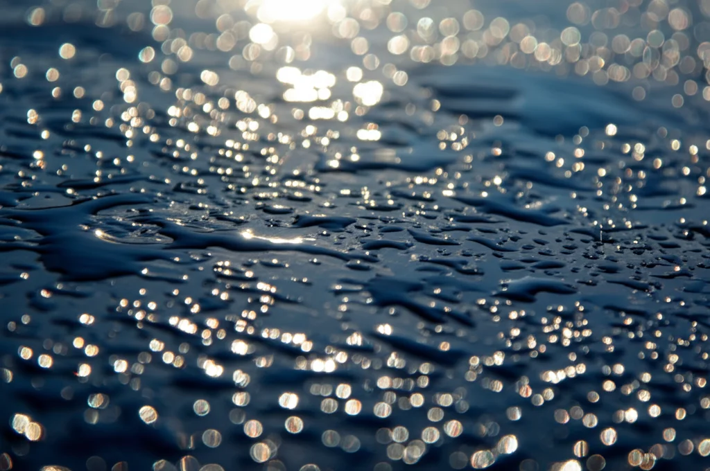 Dettaglio macro di un pannello solare termico con rivestimento nero selettivo avanzato in alluminio anodizzato, gocce d'acqua sulla superficie che riflettono intensamente il sole, lente macro 85mm, alta definizione, illuminazione naturale brillante, messa a fuoco selettiva sulla texture del rivestimento.