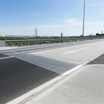 Fotografia grandangolare, 20mm, di un moderno ponte senza giunti (FJB) collegato alla strada tramite una lastra R-ECC. L'asfalto appare liscio e continuo nel punto di transizione. Cielo sereno, luce diurna, focus nitido sull'infrastruttura. Long exposure per dare un leggero effetto mosso a un'auto di passaggio.